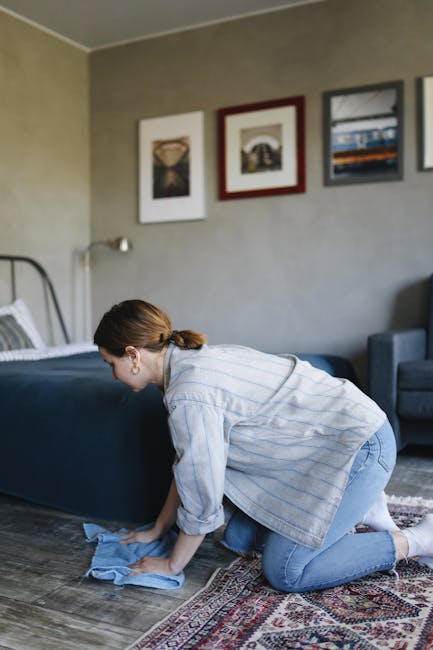 A person using a yellow vacuum cleaner with a black hose to perform surface cleaning and deep cleaning of a patterned, beige and green area rug in a living room. The individual, wearing casual clothin