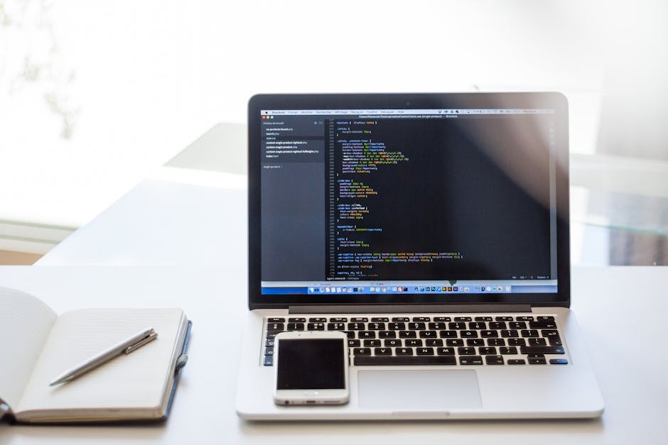 A laptop displaying a code editor with lines of programming code on a clean, white desk. Next to the laptop is a closed smartphone and a notepad with a pen resting on top. The background features a wi